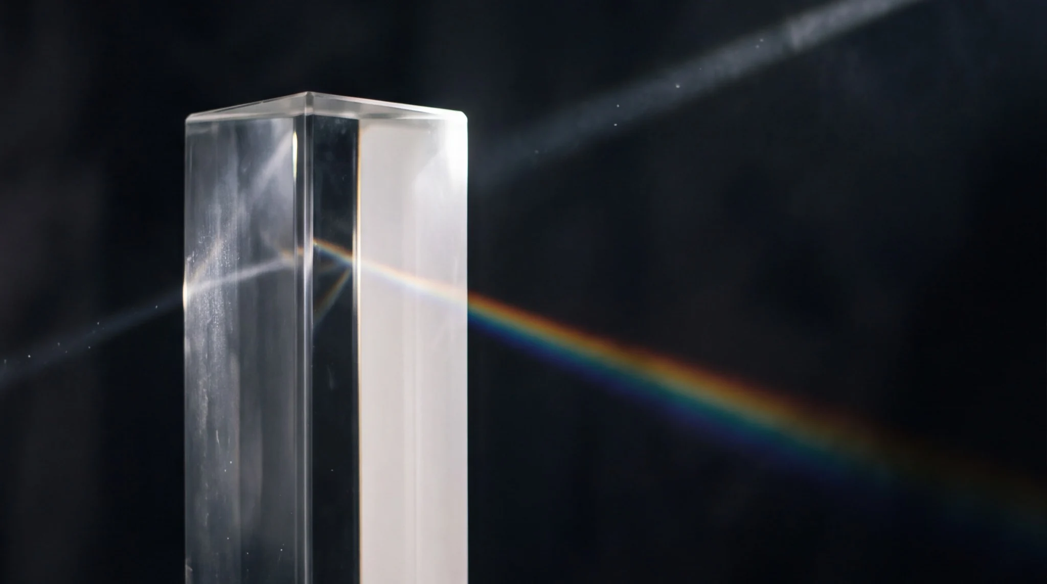 A clear rectangular prism refracts a narrow beam of white light into a subtle rainbow against a dark background.