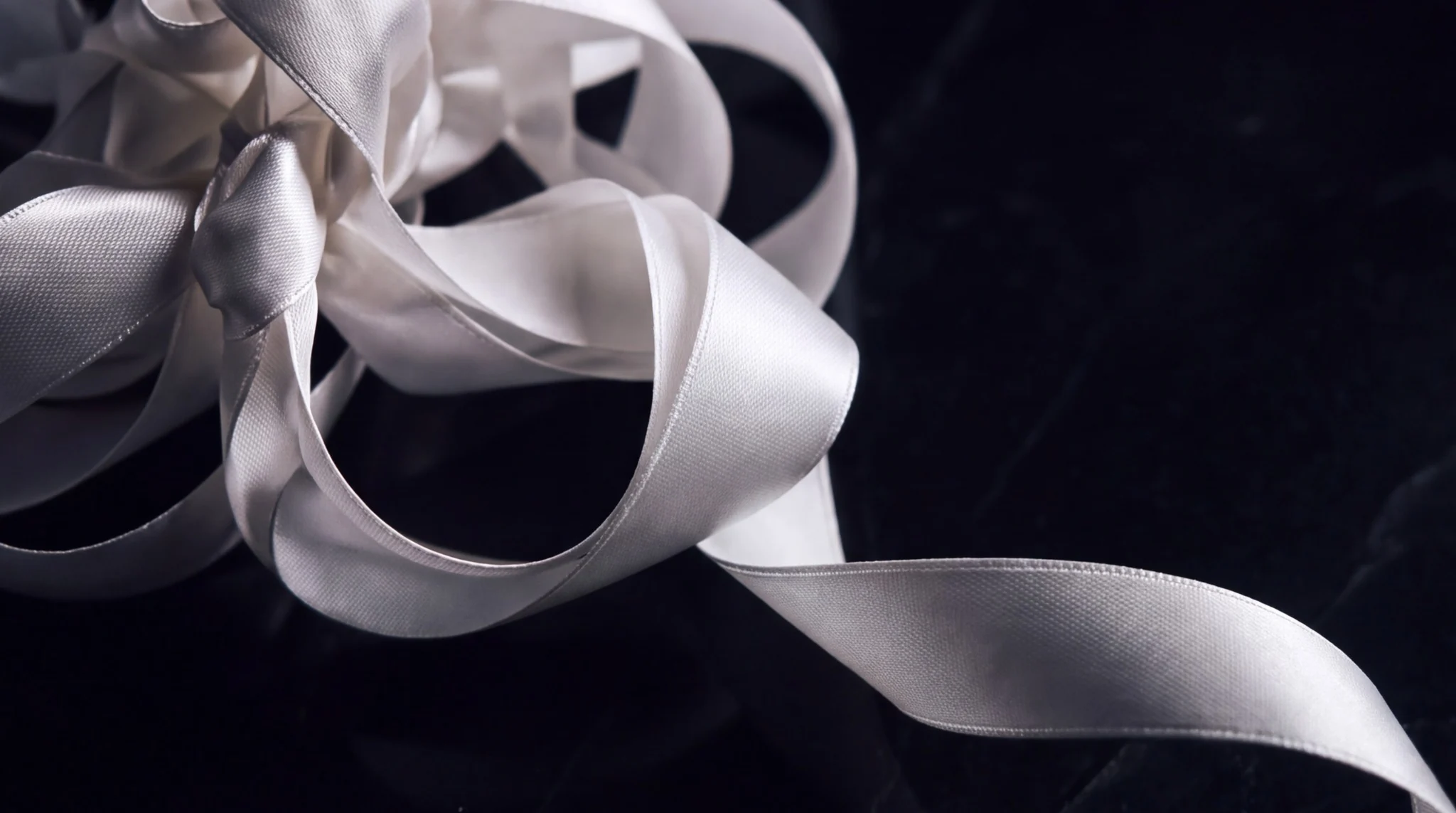 An ivory ribbon rests in loose loops on dark fabric, with one length extending away in a smooth, uninterrupted curve.